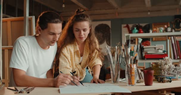 Boldog fiatal kaukázusi pár művész rajzol össze egy rajzot ceruzákkal és mosolyognak egymásra a hangulatos műhelyben. — Stock videók