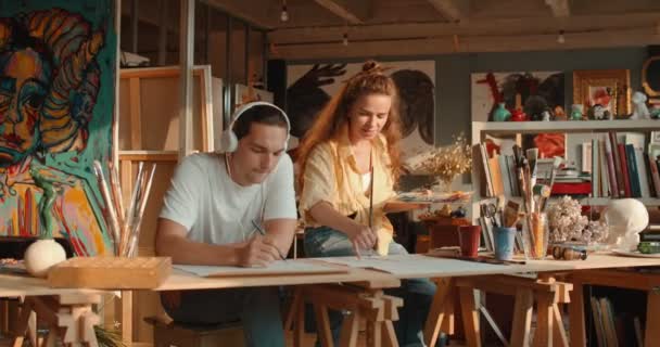 Kaukasisch jong paar kunstenaars werkzaam in de gezellige werkplaats, man die naar de muziek luistert en tekent met een potlood en vrouw schilderend met verf. — Stockvideo