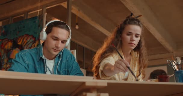 Blick unter den Schreibtisch auf die jungen kaukasischen Maler, die in der Werkstatt arbeiten und malen, Mann mit Kopfhörer, der Musik lauscht und Frau, die mit Pinsel und Farbe malt. — Stockvideo