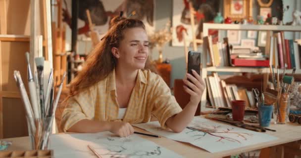Caucásica feliz joven pintora guapa joven sentada a la mesa con sus dibujos y hablando a través de webcam en su teléfono inteligente como si tuviera videochat con un amigo en el estudio de arte.. — Vídeos de Stock