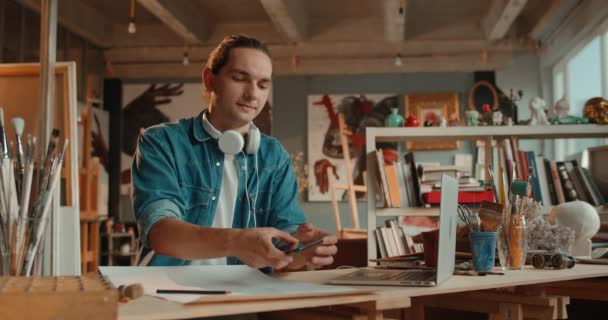 Young handsome Caucasian male student of art sitting in the artistic studio at the desk and using his smartphone, typing and chatting or playing a game. — Stock Video