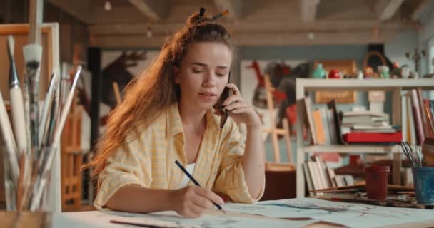 Genç Kafkasyalı yaratıcı ve stil sahibi kadın ressam atölyede çalışırken bir skeç çizip cep telefonuyla konuşuyor.. — Stok video