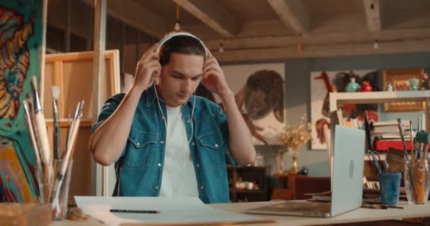 Attractive young Caucasian male artist taking on headphones and listening to the music while depicting a picture from the screen of the laptop computer while drawing with a pencil. — Stock Video