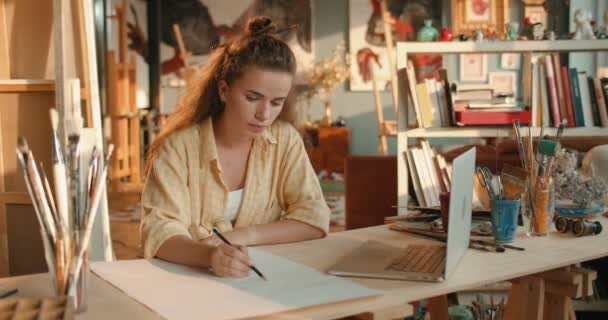 Young Caucasian pretty girl painter drawing a sketch with a pencil and depicting a picture from the screen of the laptop computer. — Stock Video