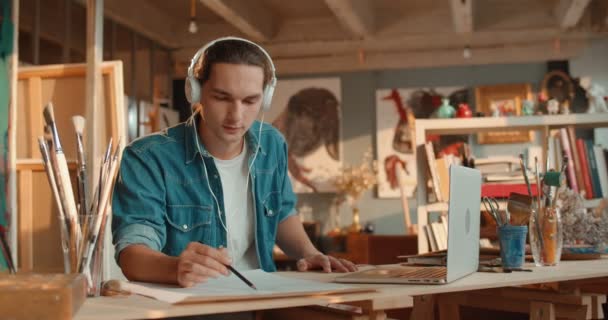 Kulaklıklı, genç, beyaz, yakışıklı bir adam. Kalemle bir eskiz çiziyor ve dizüstü bilgisayarın ekranından bir resim çiziyor.. — Stok video