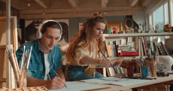 Kavkazská mladá hezká žena malovat obraz s barvami a pohledný muž ve sluchátkách poslech hudby a kreslení s tužkou na bílém listu papíru. — Stock video