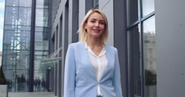 Gülümseyen ve neşeli genç sarışın beyaz kadın iş stili ile büyük şehir binasına doğru yürüdü ve kameraya baktı. Portre çekimi.