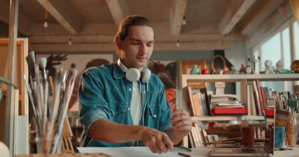 Close up of the cheerful Caucasian young guy painter sitting at the table in his working workshop and chatting on his smartphone while tapping and texting. — Stock Video