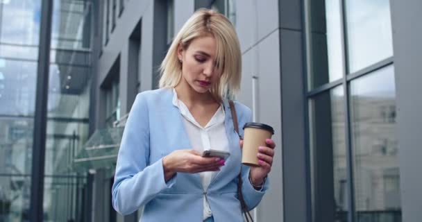 Kaukaska młoda blondynka dobrze wyglądająca stylowa bizneswoman wychodząca na zewnątrz do biura lub spotykająca się rano, pijąca kawę na wynos i stukająca lub pisząca smsy na smartfonie. — Wideo stockowe