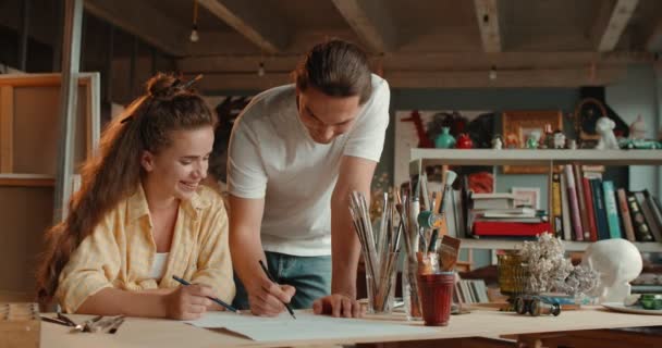 Genç, mutlu, beyaz, güzel bir kadın masada oturuyor ve bir kalemle çizim yapıyor. Öğretmen, erkek sanatçı, ona nasıl çizim yapılacağını ve öğretileceğini gösteriyor.. — Stok video
