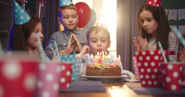 Feliz niño caucásico celebrando un cumpleaños soplando velas y sonriendo a la cámara mientras sus amigos tocan los cuernos y aplauden . — Vídeos de Stock