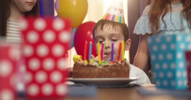 Divertido niño caucásico en cono de cumpleaños sentado delante de la torta con velas y lamiendo una crema desde la parte superior. De cerca. . — Vídeos de Stock