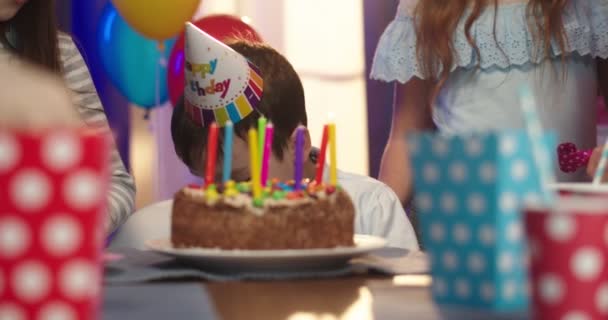 Close up of the small Caucasian cute birthday boy in conus sitting in front of the birthday cake and waiting to blow candles while hand of the mother lighting them. — Stock Video