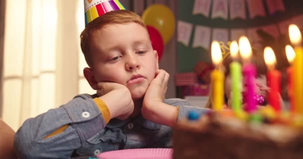 Close up do menino adolescente caucasiano entediado em um cone sentado na mesa em frente ao bolo de aniversário e soprando velas com rosto infeliz. Retrato . — Vídeo de Stock