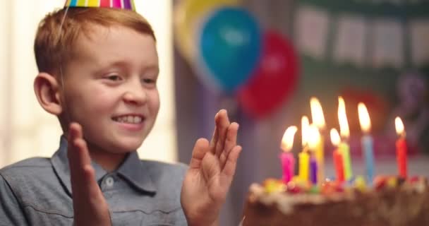 Primer plano del pequeño chico guapo caucásico en un cono celebrando su cumpleaños, soplando velas en el pastel y sonriendo. Retrato . — Vídeo de stock