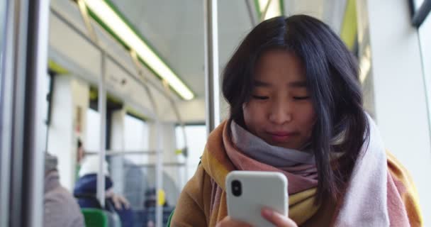 Joven chica asiática bonita escribiendo y mensajes de texto en el teléfono inteligente en el tranvía, mientras que va a alguna parte y mirando a la ventana . — Vídeo de stock