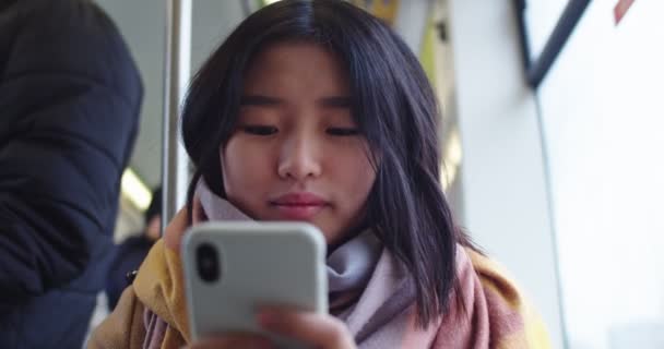 Aranyos és gyönyörű fiatal ázsiai lány kezében okostelefon és koppint rá, miközben ül a villamoson vagy a buszon. Közelről.. — Stock videók