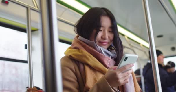 Sonrió alegre joven asiática en auriculares tocando y enviando mensajes de texto a un mensaje en el teléfono mientras escuchaba la música e iba en el tranvía o autobús. De cerca. . — Vídeo de stock