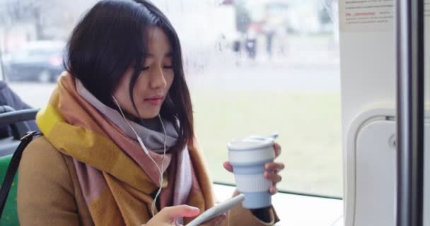 Chica joven asiática en headohones escuchando la música y tocando en el teléfono inteligente mientras toma café caliente o té y se sienta en el tranvía o autobús . — Vídeo de stock