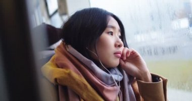 Close up of the young Asian good looking bored girl in headphones listening to the melancholic music and dreaming while looking at the window on the rainy boring day in the tram or bus.