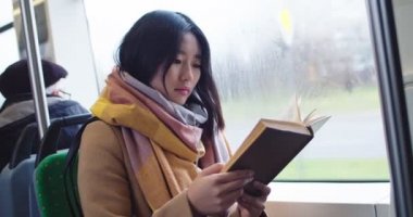 Asian young good looking girl reading interesting book while sitting in the tram or bus and going somewhere on the rainy day.