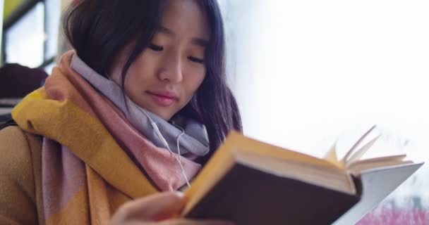 Закрийте молоду азіатську посміхнуту дівчину в навушниках, слухаючи музику, читаючи книжку або підручник, і йдіть восени на трамвай або автобус.. — стокове відео