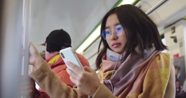 Asiática joven elegante y bonita chica en gafas y auriculares tocando en el teléfono inteligente y escuchando la música mientras está de pie en el tranvía o autobús . — Vídeo de stock
