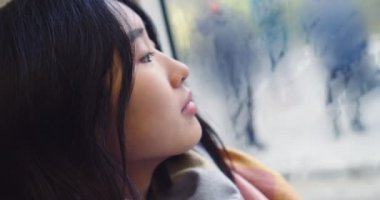 Close up of the young beautiful Asian girl looking at the window on the rainy autumn day while sitting and going somewhere in the tram or bus.