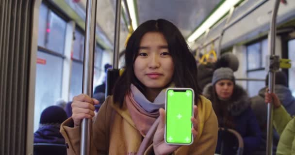 Lviv, Ucrania - 25.12.2019: Retrato de la joven asiática hermosa mujer sosteniendo un teléfono inteligente en la mano y mostrándolo a la cámara con la pantalla de croma y el movimiento de seguimiento mientras está de pie en el tranvía — Vídeo de stock