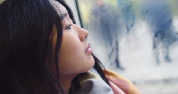 Close up of the young beautiful Asian girl looking at the window on the rainy autumn day while sitting and going somewhere in the tram or bus. — Stok video