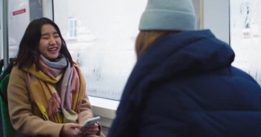 Asian young joyful girl with smartphone in hands sitting in front of her best friend in the tram or bus and talking cheerfully on a rainy day. Rear. Back view.