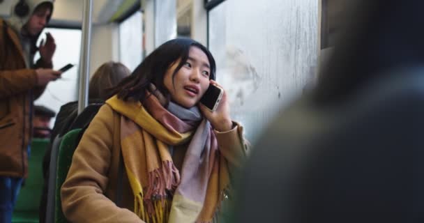 Vrolijk jong Aziatisch meisje praten aan de telefoon en glimlachen terwijl zitten aan het raam in de tram of bus op een regenachtige dag. — Stockvideo