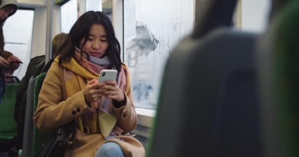 Stylish young Asian girl tapping and texting on the smartphone while sitting at the window in the tram or bus on a rainy day. — 图库视频影像