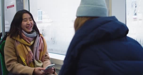 Chica joven asiática alegre con teléfono inteligente en manos sentadas frente a su mejor amiga en el tranvía o autobús y hablando alegremente en un día lluvioso. Atrás. Vista trasera . — Vídeo de stock