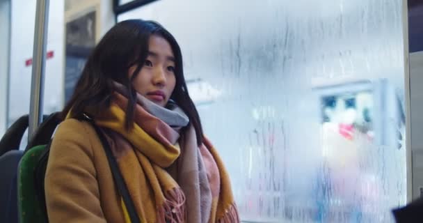 Młoda piękna azjatycka kobieta w stylowym stroju z szalikiem siedzącym w tramwaju lub autobusie przy oknie, idąc gdzieś i wycierając szkło z wilgotności. — Wideo stockowe