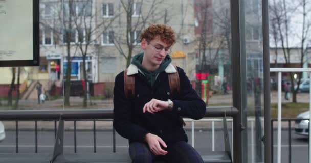 Caucasian young guy in glasses and with red hair sitting on the tram or bus stop, looking at the watch and waiting for the transport. Outdoor. — 图库视频影像