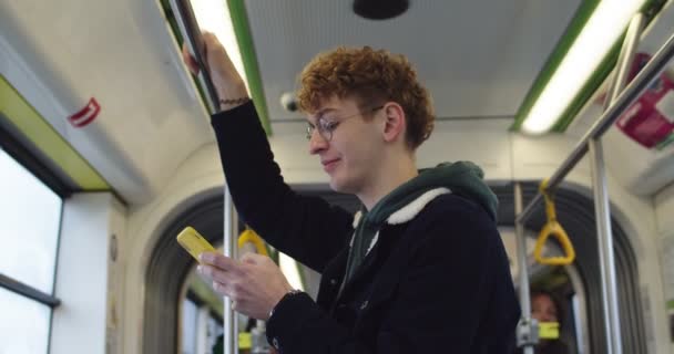 Joven chico caucásico de pie en el tranvía o autobús como ir a alguna parte, tocando y mensajes de texto en su teléfono inteligente . — Vídeos de Stock