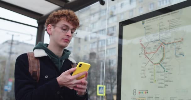 Young Caucasian red-haired guy in glasses standing at the tram or bus stop and tapping or scrolling on the smartphone while waiting fot the transport. — Stok video