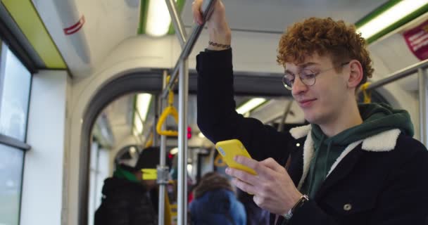 Caucasian young guy in glasses and with red hair texting a messsage while chatting on the smartphone in the tram or bus and smiling. — 图库视频影像