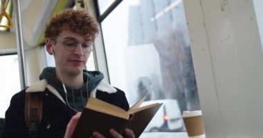 Caucasian young guy in glasses, headphones and with red hair reading interesting book while sitting in the tram or bus and going somewhere on the rainy day.
