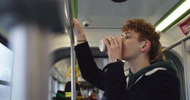 Chico pelirrojo caucásico feliz y sonriente con gafas de pie en el tranvía o autobús y bebiendo té caliente o café para llevar mientras va a alguna parte . — Vídeo de stock