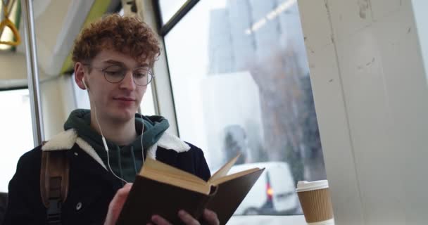 Blanc jeune homme en lunettes, écouteurs et avec les cheveux roux lecture livre intéressant tout en étant assis dans le tram ou le bus et aller quelque part le jour de pluie . — Video