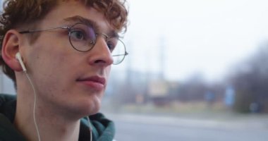 Close up of the young Caucasian guy in glasses and with red hair listening to the music in headphones while sitting in the tram or bus at the window.