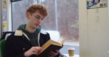 Red-haired Caucasian young guy in glasses reading a book or textbook while sitting in the tram or bus at the window.