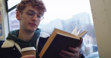 Caucasian young red-haired guy in glasses sitting at the window at the tram or bus and reading interesting book while going somewhere and sipping hot tea or coffee to-go. Close up.