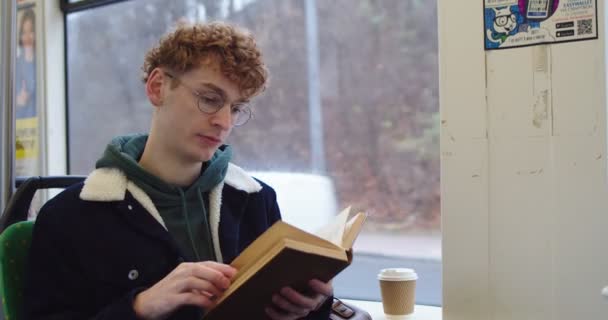 Roodharige blanke jongeman met bril die een boek of leerboek leest terwijl hij in de tram of bus aan het raam zit. — Stockvideo