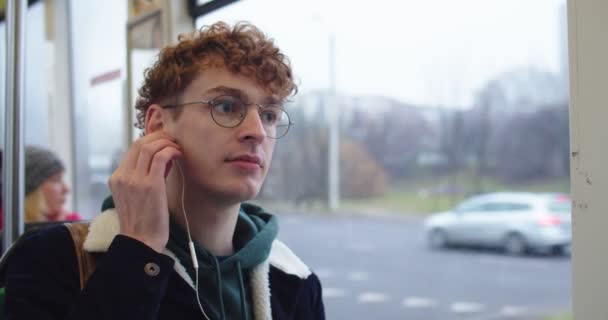 Blanc jeune homme aux cheveux roux en lunettes mettre des écouteurs blancs dans ses oreilles et écouter de la musique tout en allant quelque part dans le tram ou le bus . — Video