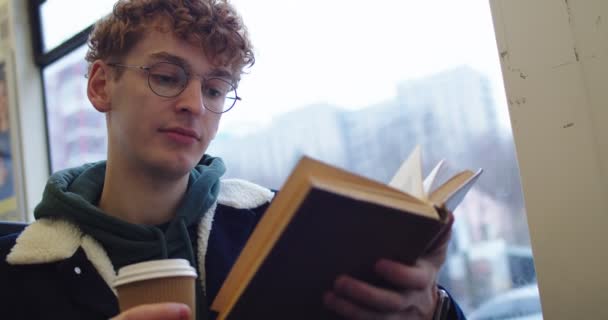 Caucasian young red-haired guy in glasses sitting at the window at the tram or bus and reading interesting book while going somewhere and sipping hot tea or coffee to-go. Close up. — 비디오