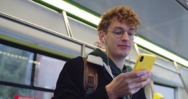 Young red-haird Caucasian guy in glasses and headphones tapping and texting on the smartphone while listening to te music and going somewhere in the tram or bus.