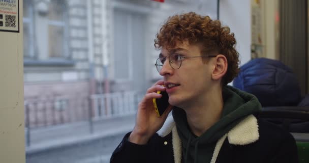 Καυκάσιος νεαρός φοιτητής με γυαλιά και με κόκκινα μαλλιά μιλώντας στο κινητό τηλέφωνο ενώ κάθεται στο παράθυρο στο τραμ ή το λεωφορείο και πηγαίνει κάπου. — Αρχείο Βίντεο